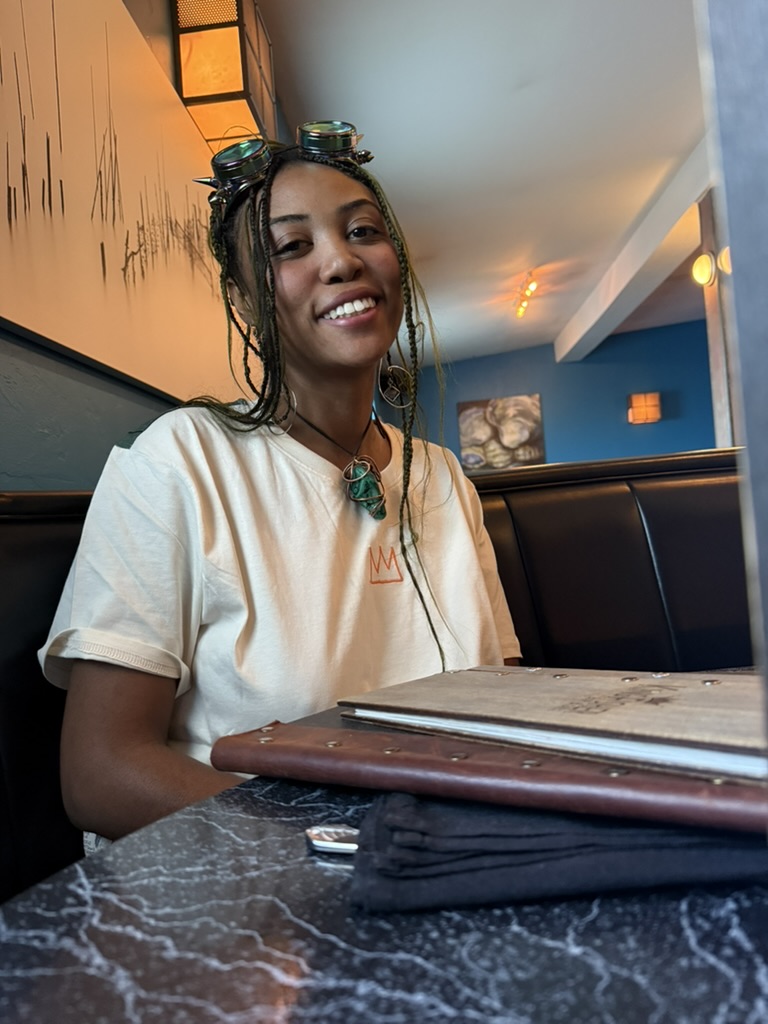 Warm natural-light portrait of Kayla Janae, a smiling young Black woman with braided hair wearing a mustard yellow top, standing in front of a colorful painted mural.